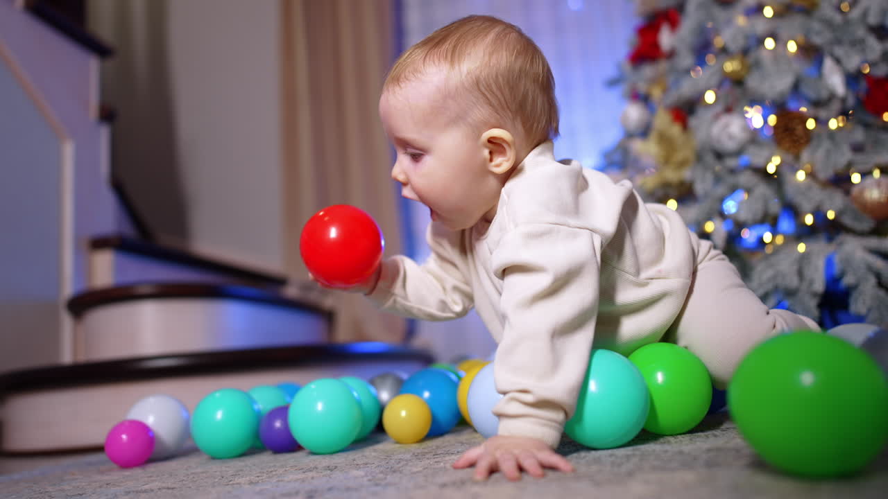 Beautiful baby in white sport suit crawls by the floor. Cute child picks the plastic balls scattered around him. Close up. Christmas tree at backdrop in blur.