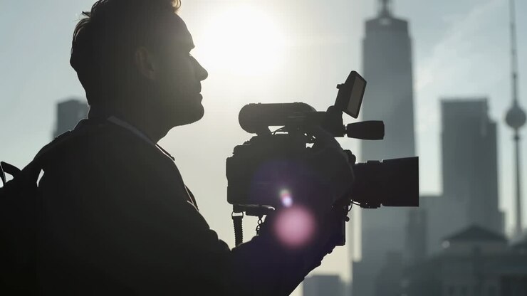Videomaker uomo che regola l'inquadratura con una spalliera su un tetto sopra la città, contrastando il bagliore solare.