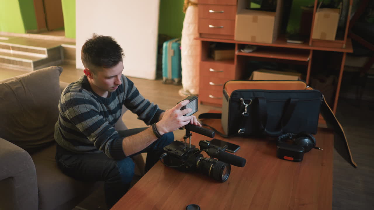 Person reaching into camera bag, pulling out equipment like lens and microphone. Preparing for filming in home studio, with camera setup and tripod ready for action