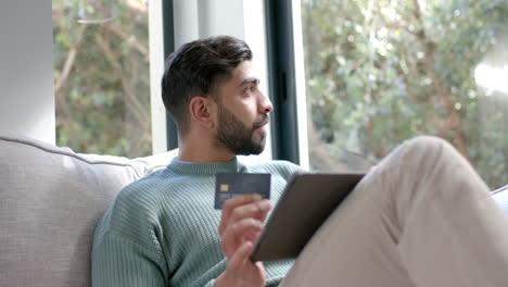 Hombre-Birracial-Sentado-En-Un-Sofá-Usando-Una-Tableta-Para-Comprar-En-Línea-En-Casa,-Cámara-Lenta