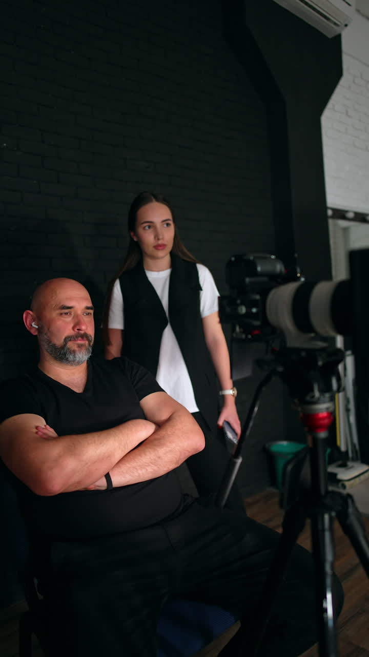 Bearded man in black clothes sits on the chair pointing at his camera. Brunette woman stands behind looking at equipment and nodding. Studio backstage. Vertical video.