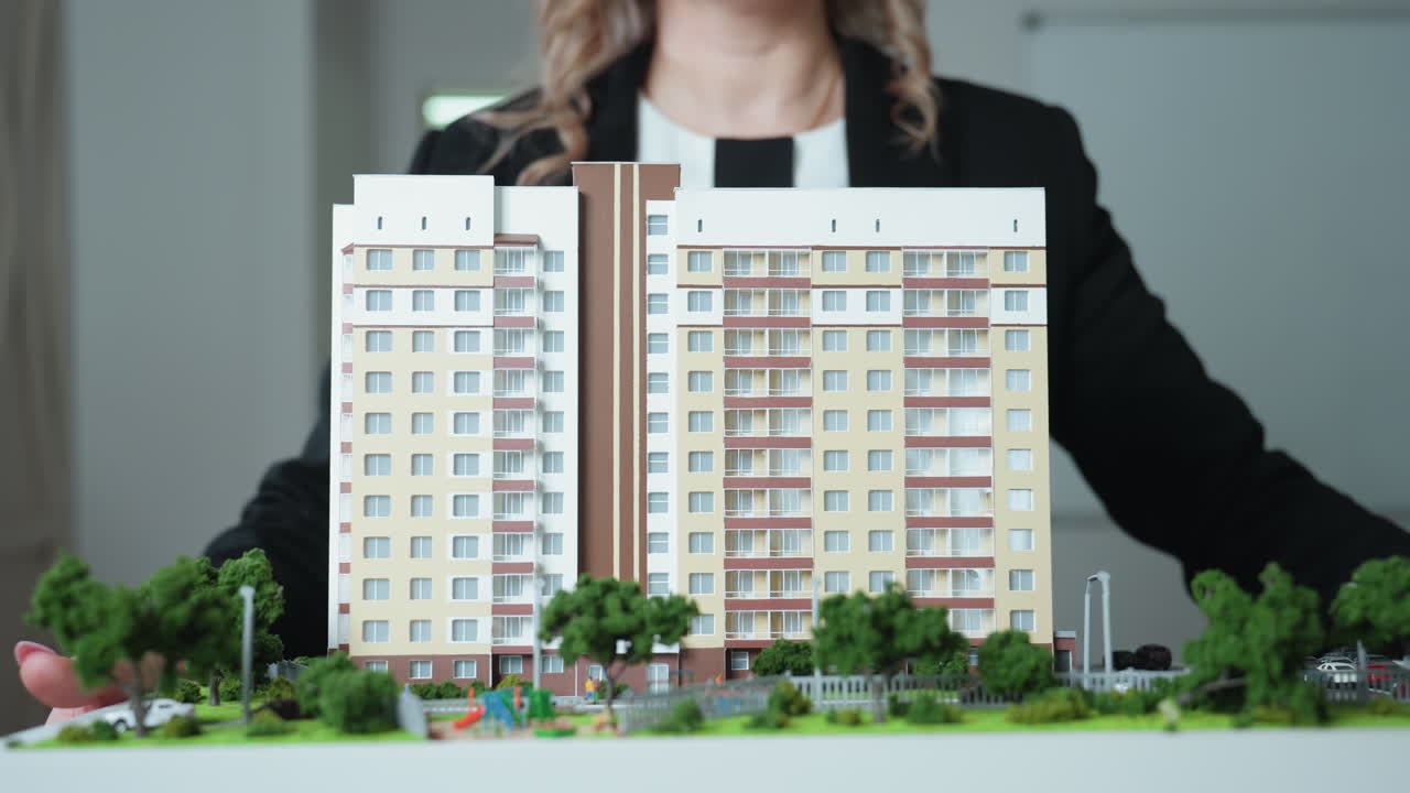 Close up of professional woman dressed in black carrying detailed building model toward office desk, showcasing modern real estate concept and architectural miniature with urban layouts
