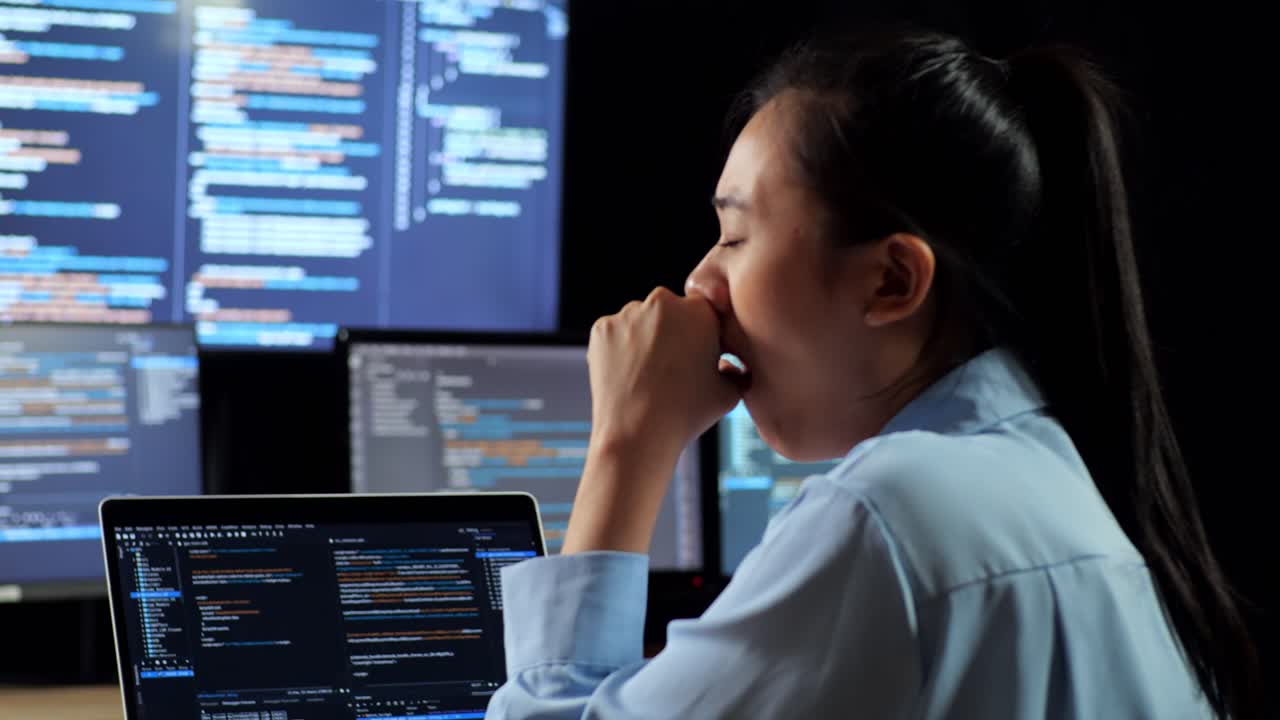 Close Up Back View Of Asian Female Programmer Yawning And Sleeping While Writing Code By A Laptop Using Multiple Monitors Showing Database On Terminal Window Desktops In The Office