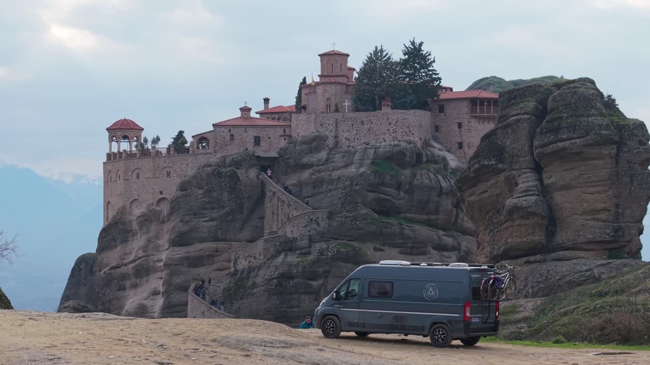 Scenic drone shot of a campervan parked on a hill at Meteora, Greece. A majestic monastery is revealed in the background, blending travel freedom with ancient heritage in an autumn landscape