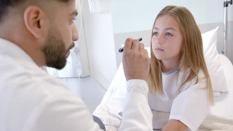 diverse male doctor checking eyes of girl patient in hospital bed with penlight, slow motion