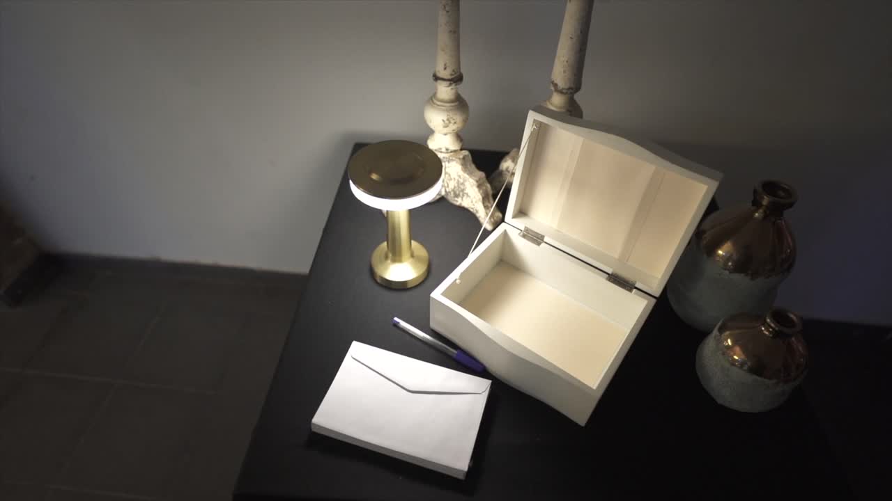 Camera slow spin over hite message box sits open on a black tabletop beside a blue pen and envelope, surrounded by candlesticks, vases, and a lamp, creating a refined and welcoming guest station