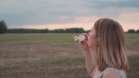 side view of young fair skinned woman in denim dress holding bubble wand and blowing bubbles along roadside near green field with distant trees under soft sunset light