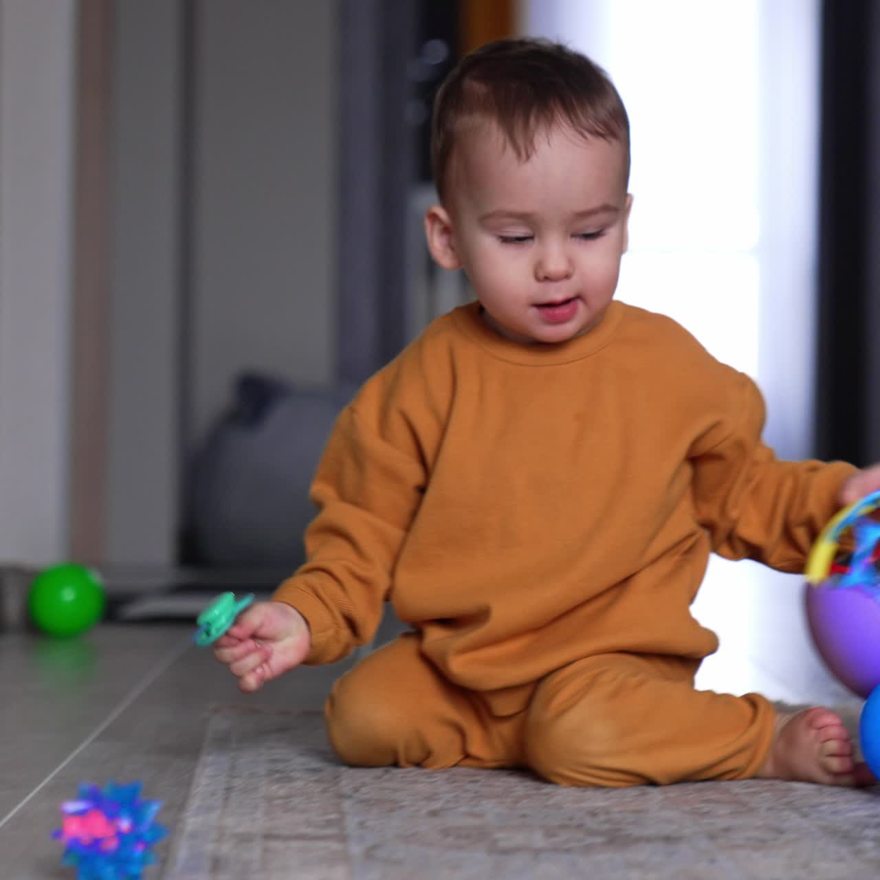 Cute kid with pacifier in his hand sits on the floor looking at the ball. Little toddler approaches the toy and pushes it, watching it rolling