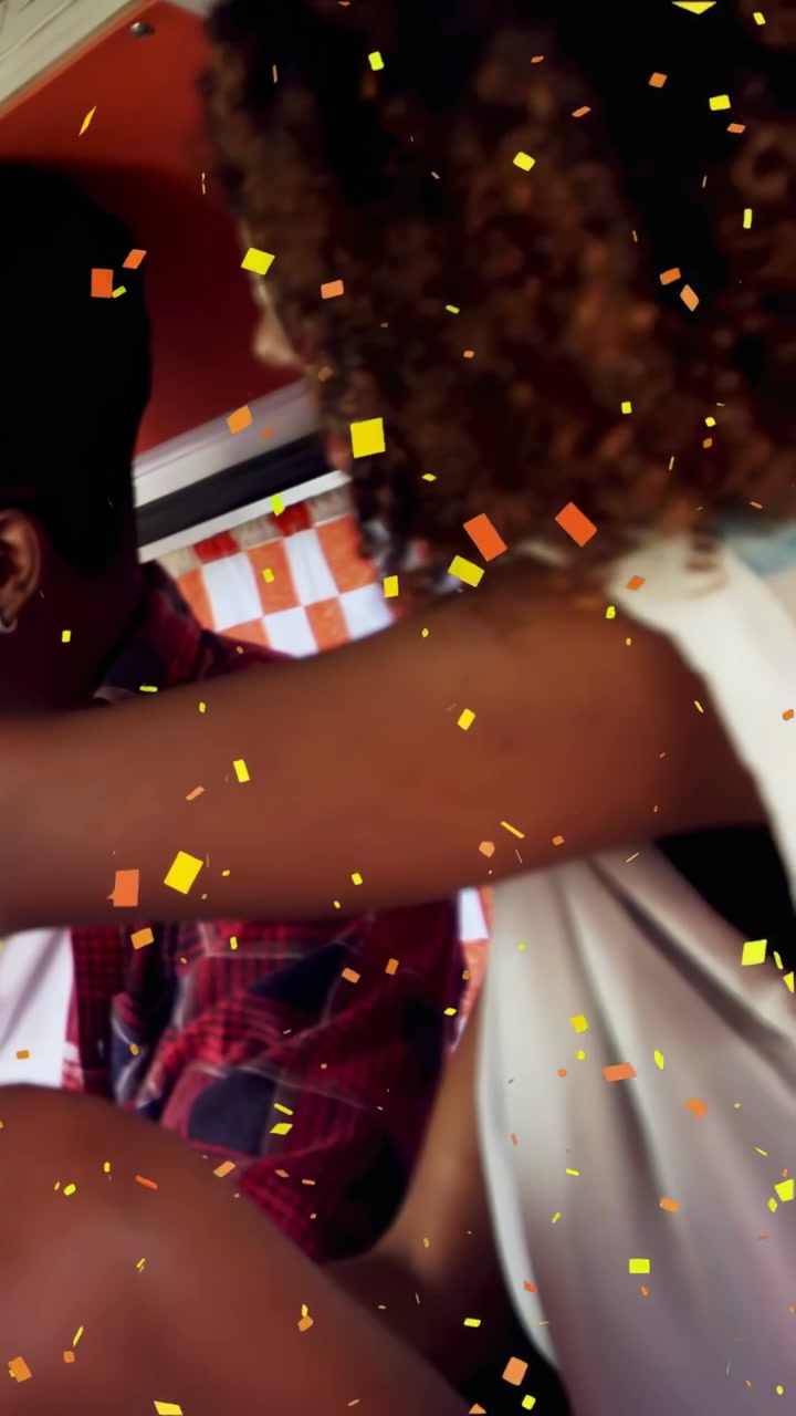 Vertical video: Woman leaning across booth reaching to grab item near man, confetti overlay for ads