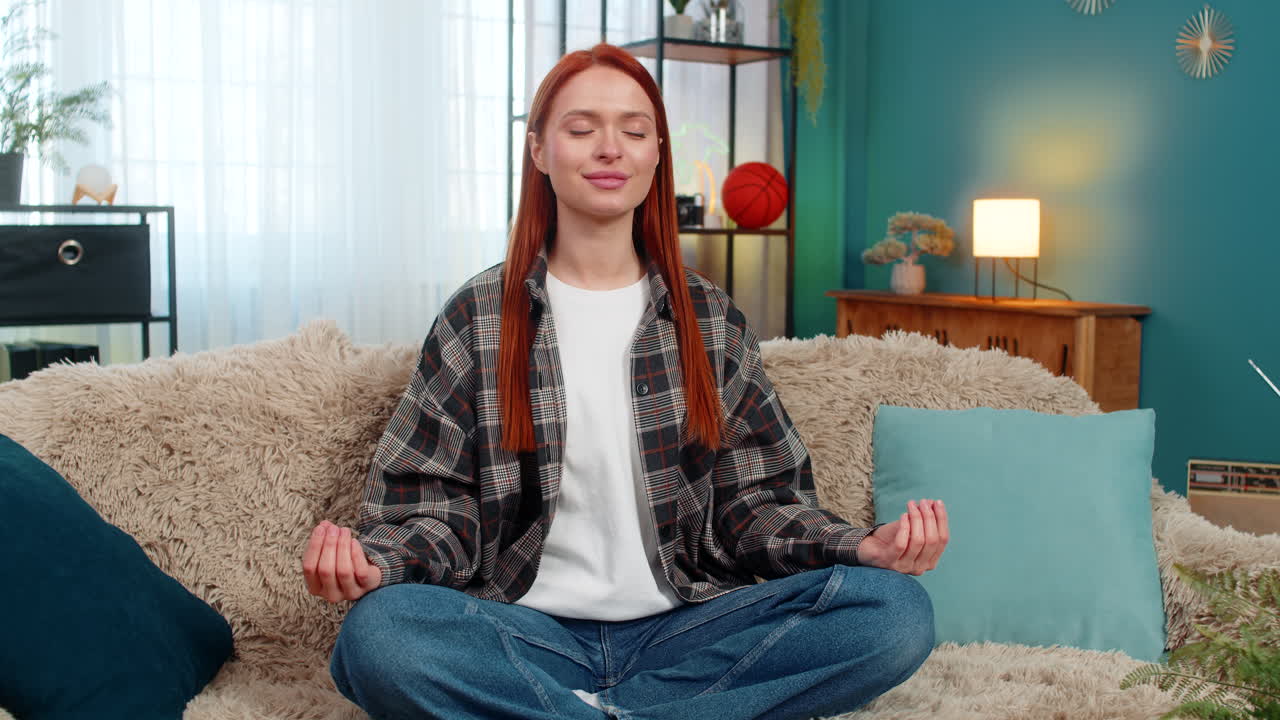 Woman sitting on sofa in lotus pose meditating with closed eyes breathing deeply feeling inner peace