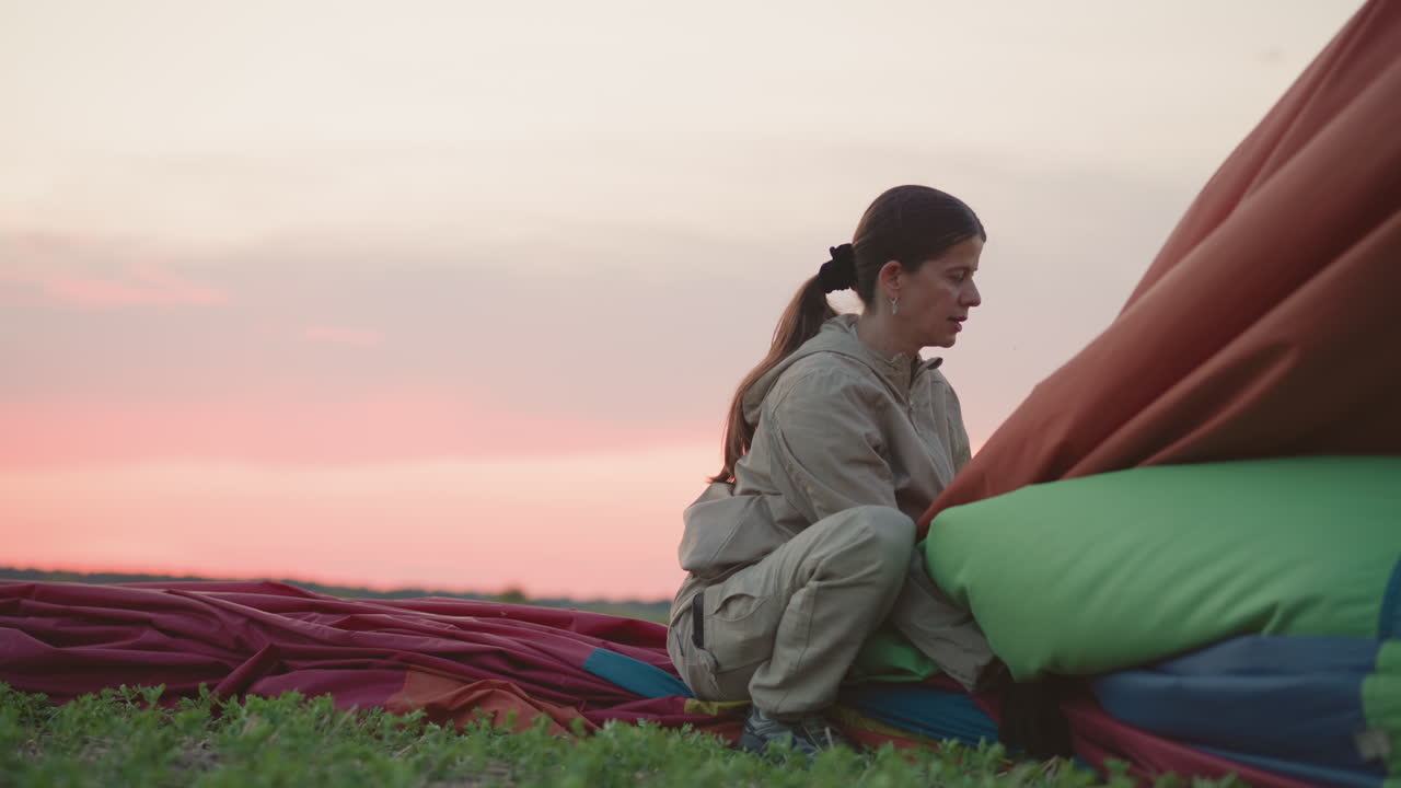 woman kneeling on deflated hot air balloon canopy on grassy field at sunset pulling fabric panel into basket near blurred passerby silhouette under soft pastel sky during twilight landing sequence