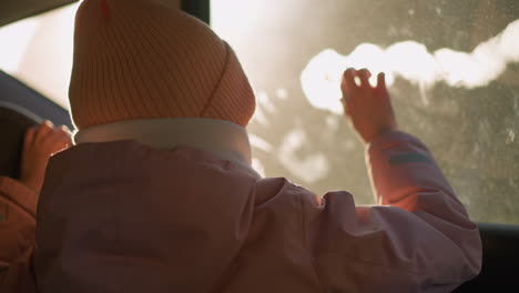 a little girl in a pink cap, jacket, and white winter scarf sits in a car, playfully writing on the fogged window. the backlit sunlight highlights her hands and the window