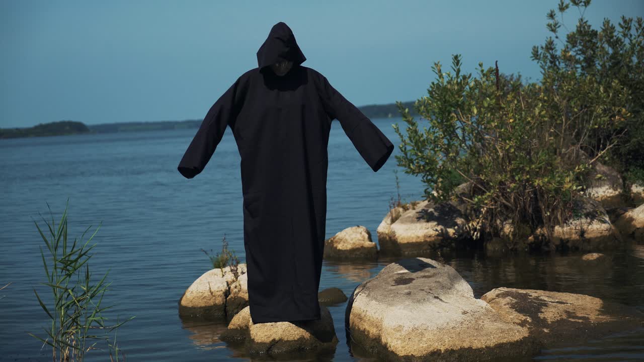 Woman devil in black cape and hood on stones near the river. Halloween costume.
