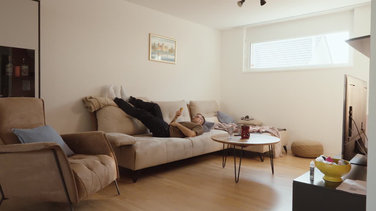 Plano aberto de um homem deitado em um sofá olhando para o seu smartphone com uma expressão de tédio, enquanto a câmera se aproxima lentamente durante a cena, mostrando fadiga digital, relaxamento e a vida doméstica cotidiana.