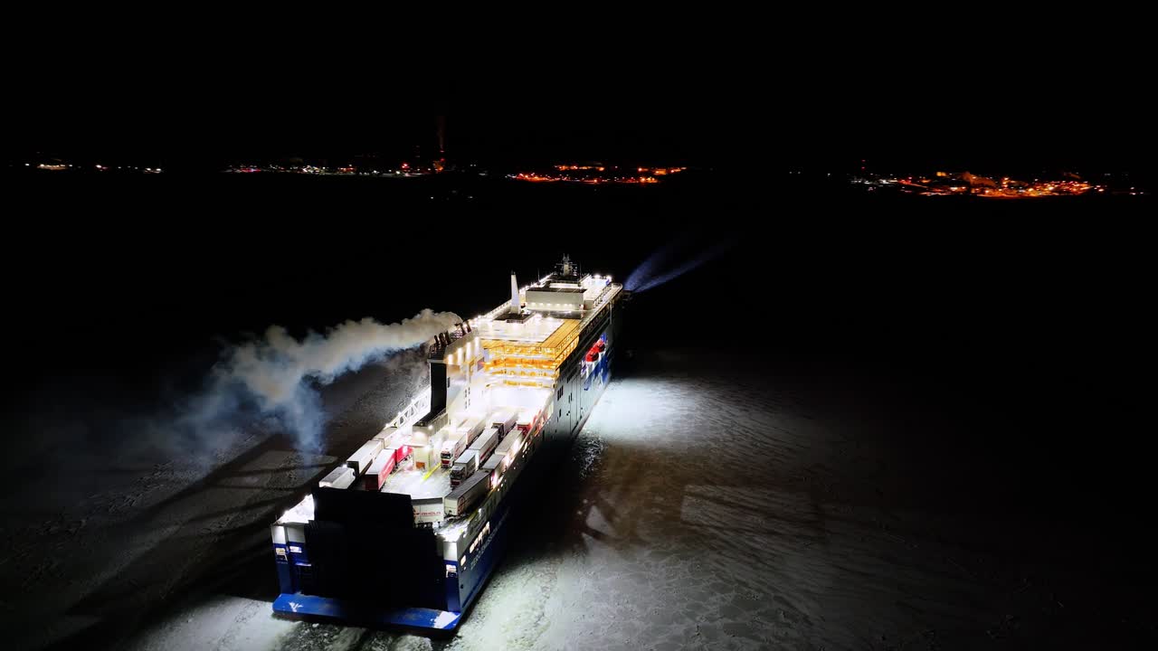 Finnlines hybrid roro ship FINNSIRIUS moving through ice covered sea at night. Searchlight illuminating ice in front. Harbour lights ahead. Aerial view crossing stern.