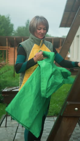 mujer de mediana edad en chaqueta amarilla y verde con botas amarillas se encuentra cerca del banco de columpio, desplegando un impermeable verde, el fondo presenta vegetación exuberante, árboles y jardín colorido