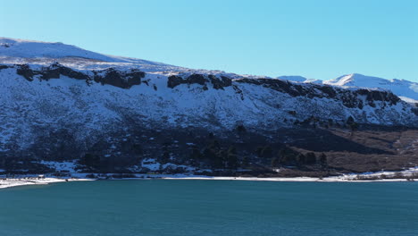 Left-moving aerial across Lake Caviahue in Neuquen, Patagonia, with turquoise water, snow capped Andean ridges and dark basalt cliffs in the Copahue volcanic landscape, smooth left pan drone pass