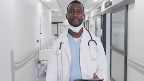 happy african american male doctor with face mask using tablet in hospital corridor, slow motion