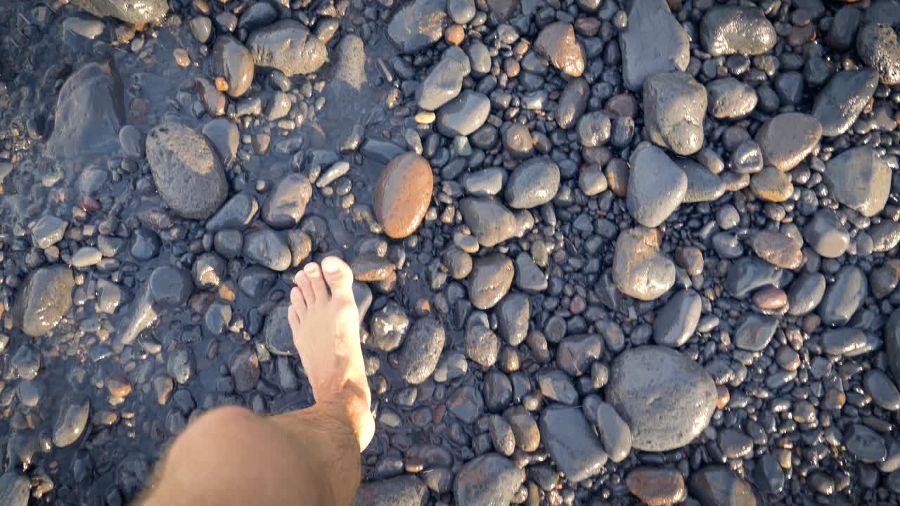 Bare feet walking on beach covered in smooth stones as waves wash over and splash. Person walking along the shore with clear warm water rushing in from left. Look down.