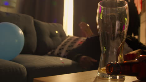 man lying on sofa after drinking alcohol at house party suffering with hangover with glasses and bottles in foreground