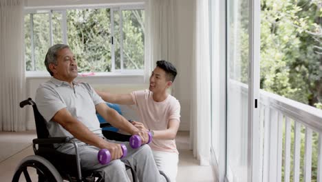 diverse male physiotherapist advising and senior man in wheelchair using dumbbells, in slow motion