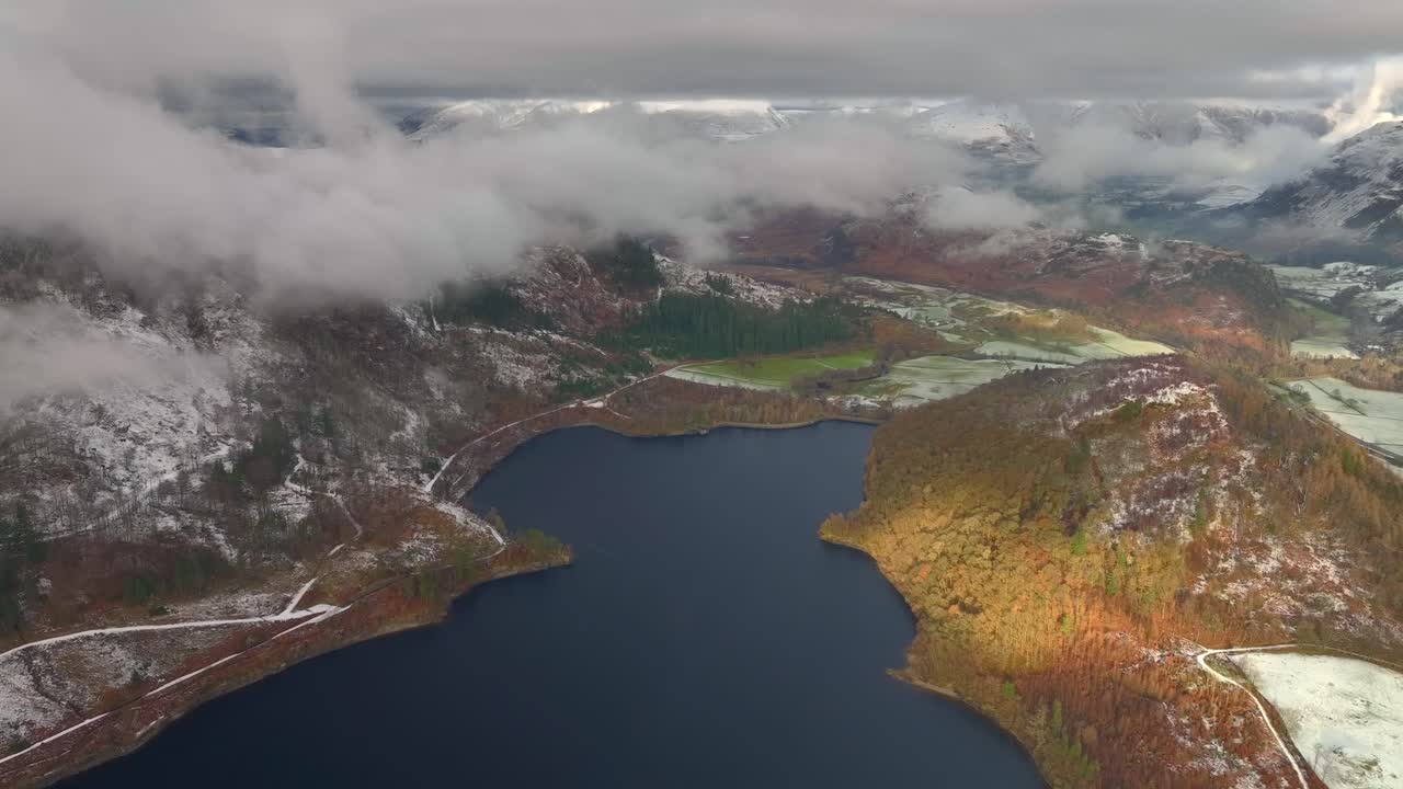 Lago oscuro y montañas con nieve ligera y un rayo de sol matutino en la ladera arbolada.