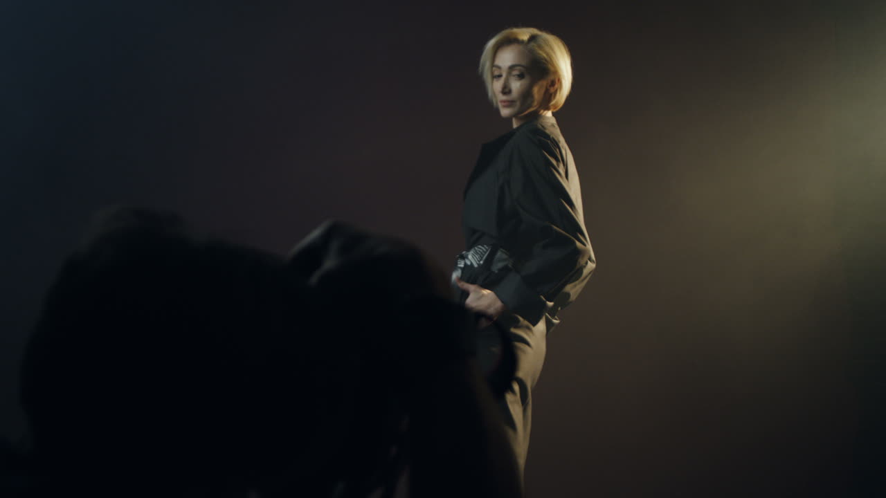 어두운 벽 배경에서 스포트라이트를 받으며 백인 여성 모델에게 사진을 찍는 사진사의 뒷모습