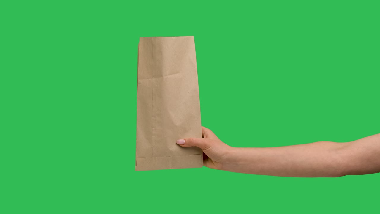 la mano femenina sostiene una bolsa de compras de papel en el fondo de una llave de croma de pantalla verde. primer plano de embalaje de papel. concepto de servicio de mensajería de alimentos y bebidas. publicidad. cámara lenta