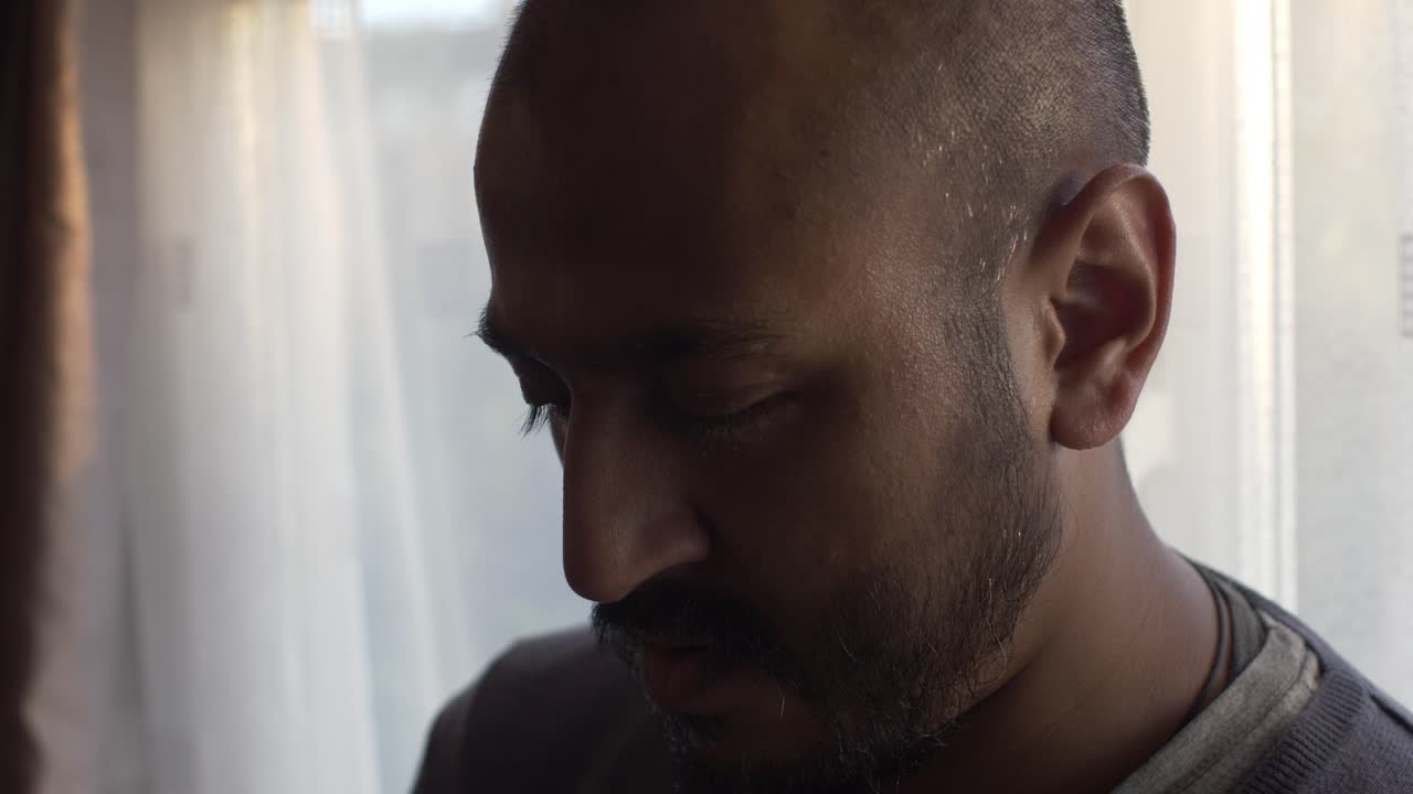 Homem asiático com expressão abatida perto de uma janela com luz de fundo e cortinas brancas