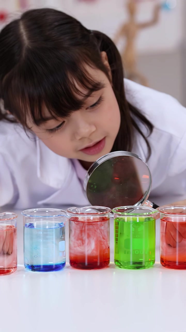 Young student in lab coat uses magnifying glass to inspect colorful liquids in glass beakers.