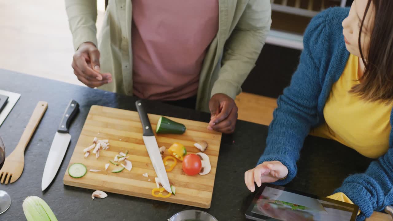 video de una feliz pareja diversa preparando comida usando la receta en la tableta en la cocina en casa