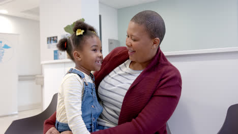 happy african american mother holding daughter on knees in hospital waiting room, slow motion