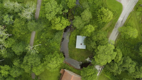 Aerial-view-over-a-car-on-a-road,-river-and-a-small-waterfall-under-a-bridge,-cloudy,-summer-day,-in-Fagervik-village,-Inkoo,-Finland---top-down,-drone-shot