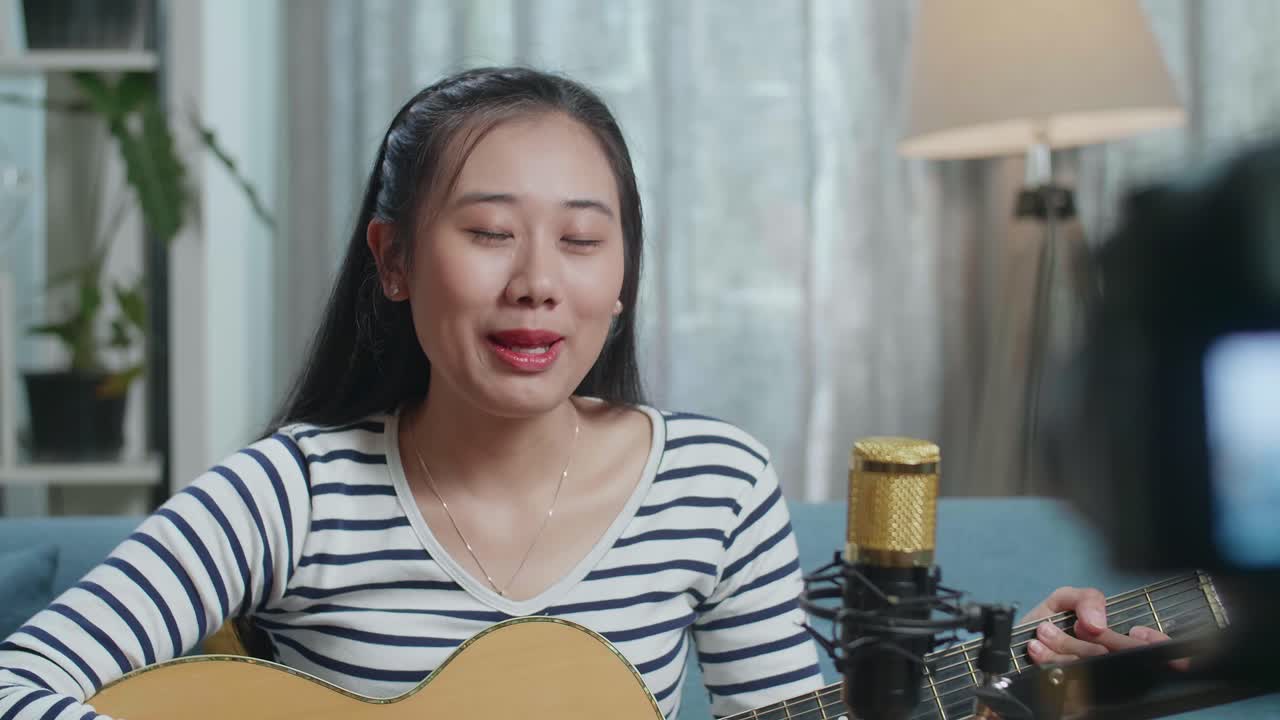 아시아 여성이 집에서 기타를 연주하는 라이브 스트리밍을 하는 동안 손을 흔들고 카메라에 말하고 있습니다.