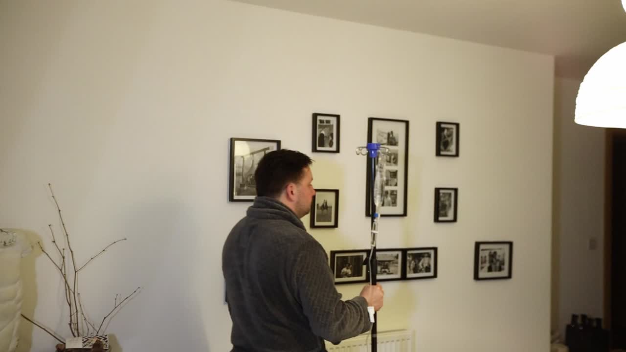 천천히 거실을 걷고 있는 남성 가정 환자가 포도당 iv 드립 백을 밀고 있습니다.