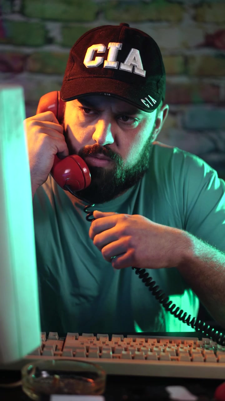 Vertical View, CIA Agent at Desk Talking Over Landline Telephone In Front of Desktop Computer From 1990's