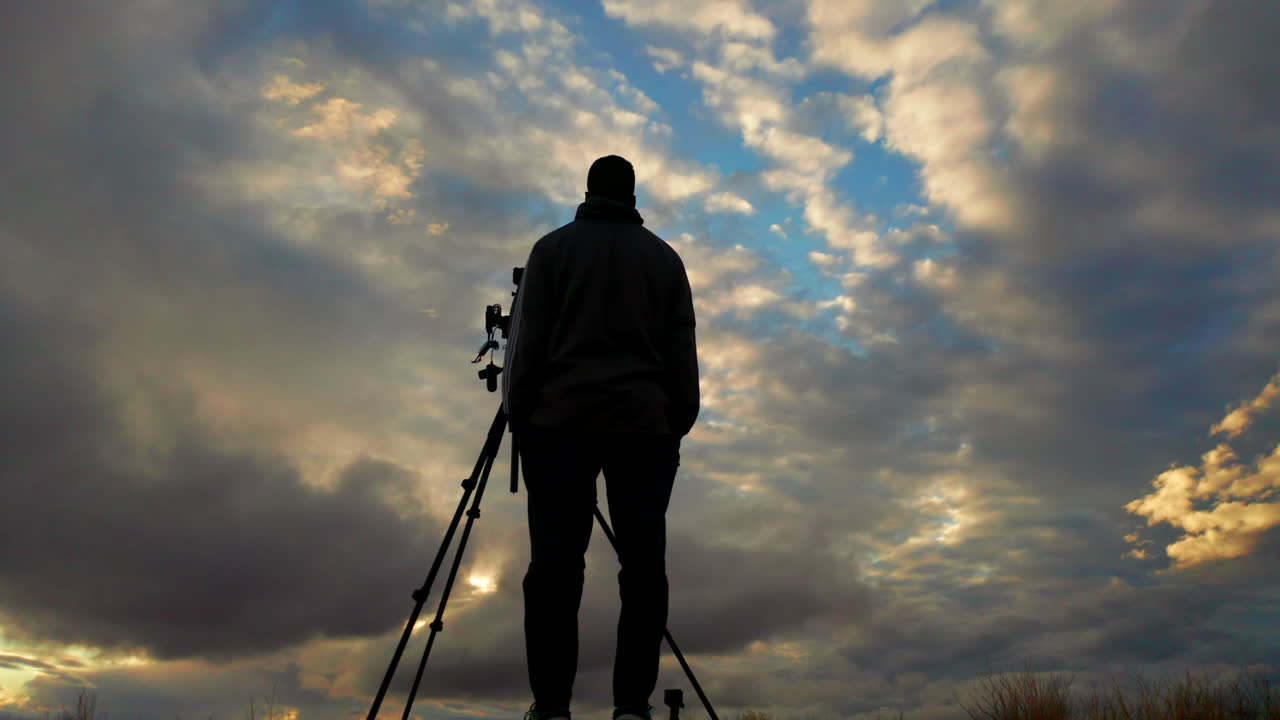Siluettfotograf som kontrollerar kameran på stativ mot en molnig himmel.