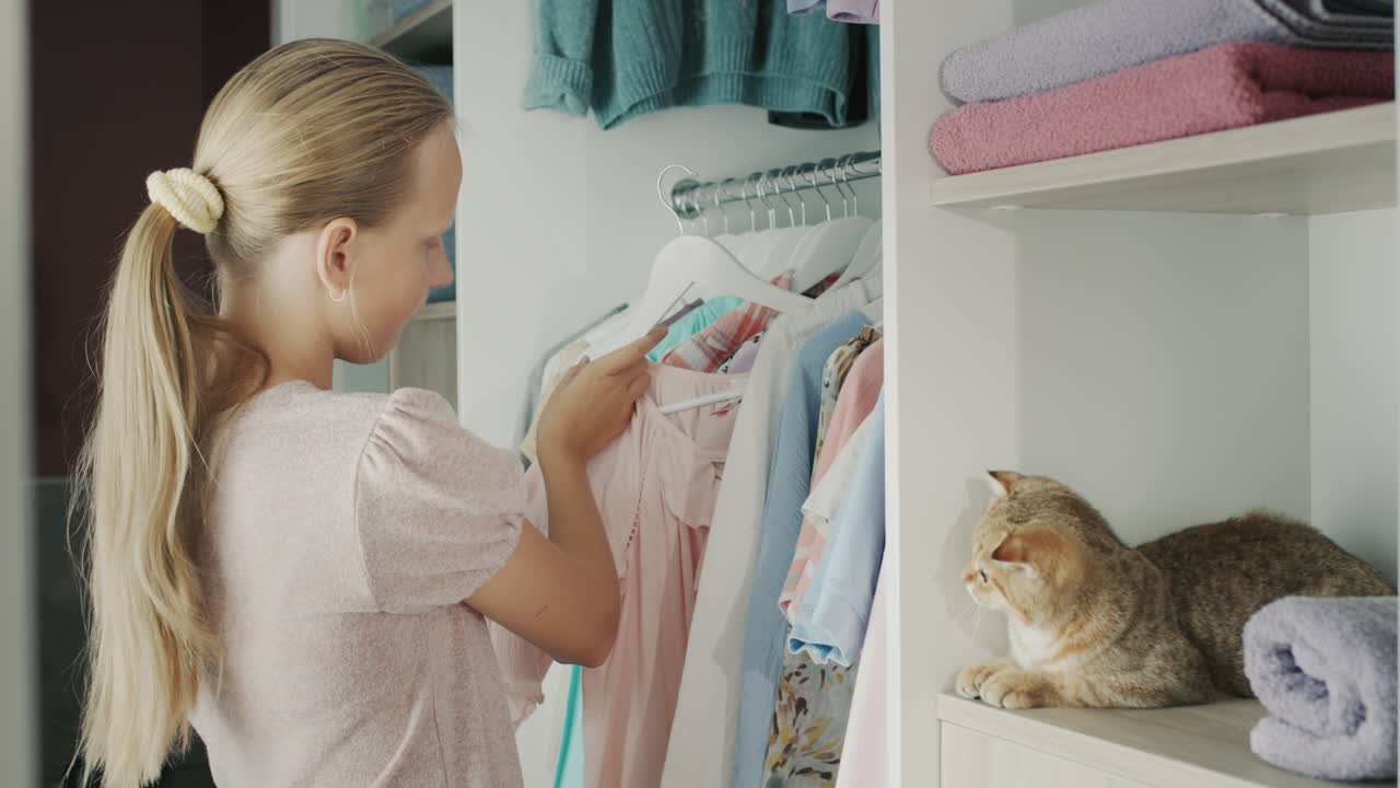 십대는 어떤 드레스를 입을지 선택하고, 그의 드레싱  ⁇ 에 있는 옷을 봅니다. 빨간 고양이가  ⁇ 에 앉아 있습니다.