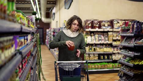 Front-view-of-caucasian-woman-walk-with-a-cart-near-shot-shelves-choosing-a-glass-jar-in-grocery-market-and-put-it-to-the-cart