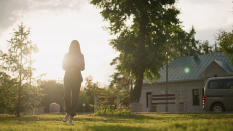 lady in grey clothing walking on grassy field outdoors during golden hour while reading a book, sunlight illuminates the surroundings, with a view of a parked car and trees