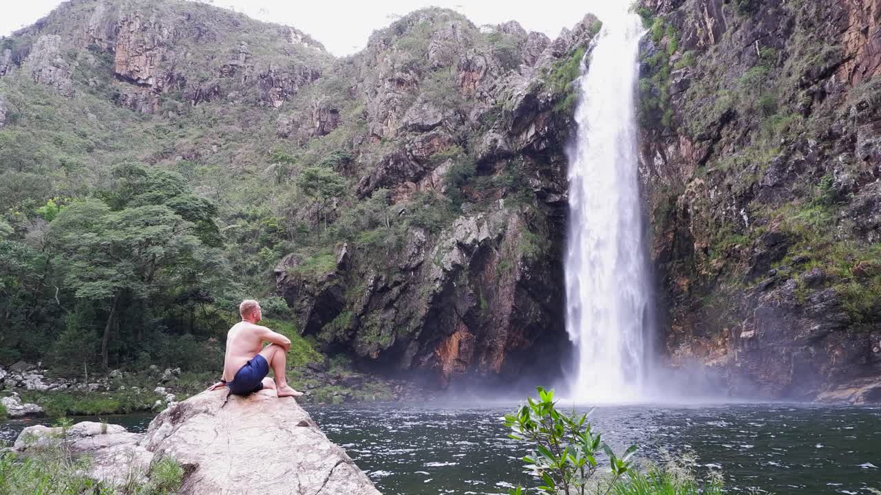 Young Caucasian man in swim trunks sits on rock below waterfall