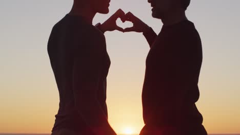 happy caucasian gay male couple holding hands and making heart shape at sunset on the beach