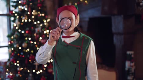 elf man with magnifying glass inside the decorated room. surprised elf is looking through the magnifying glass on christmas tree background.