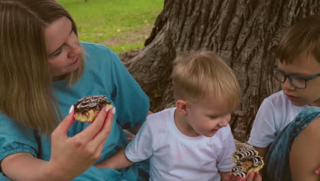 family seated under a large tree, the younger child excitedly opens his mouth to take a playful bite of his snack, while the mother watches on, holding another snack