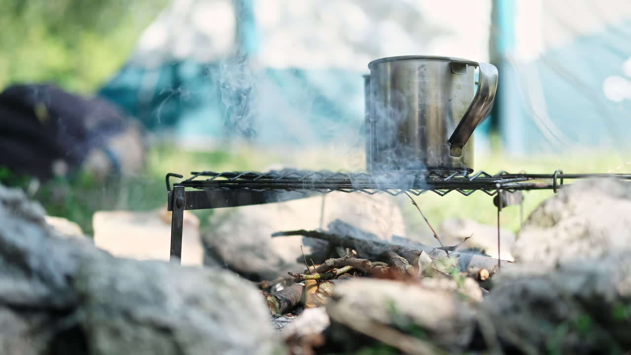 Water in two tin mugs is boiling on a grill grid above the campfire. Man is adding wood. Preparing coffee