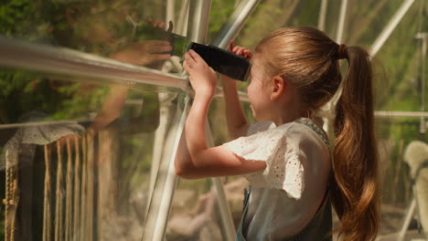 child looks in camera lens in glamping cabin. little girl records video through glass wall on sunny summer day. kid shots view of eco camping