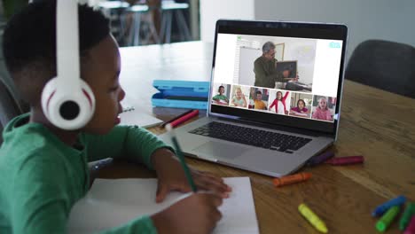 Schoolboy-using-laptop-for-online-lesson-at-home,-with-diverse-teacher-and-class-on-screen