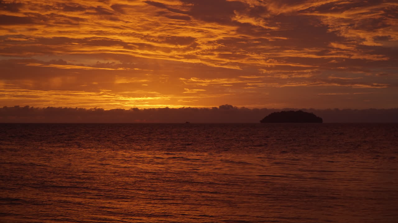 Golden sunset on a beach - Sun hides behind dence clouds, vibrant orange red sunlight reflected in sky and sea water, silhouetted islet in distance - static slow motion