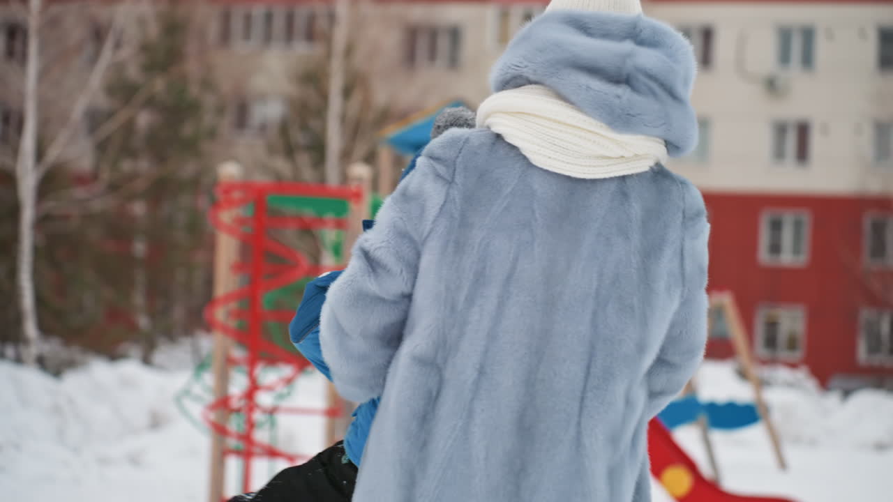 Karda oğluna bakan, örgü şapkasını ve atkısını düzelten, mavi suni kürklü palto giymiş beyaz tenli anne, arka planda renkli bir kaydırak ve tırmanma oyuncağı, apartman bloğu ve donmuş ağaçlar. Nazik koruyucu hareketler, sıcak bir aile ortamı.