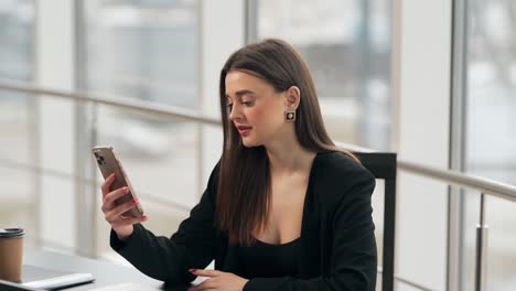 Mid-plan-of-a-young-attractive-girl-in-a-business-suit-sitting-at-a-table-with-a-smartphone